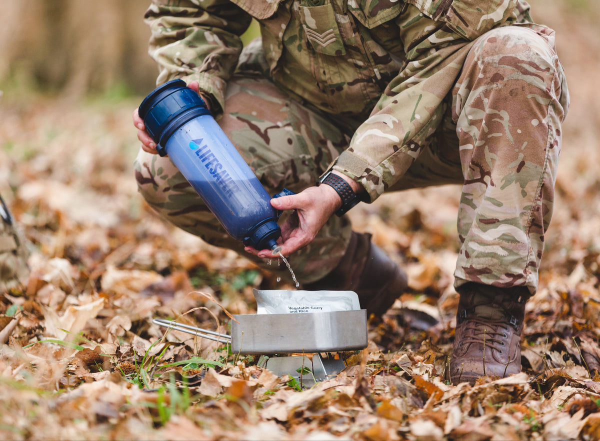 A person in camouflage military clothing crouches outdoors on a leaf-covered ground, holding a blue Lifewater water filter bottle above a metal cup.